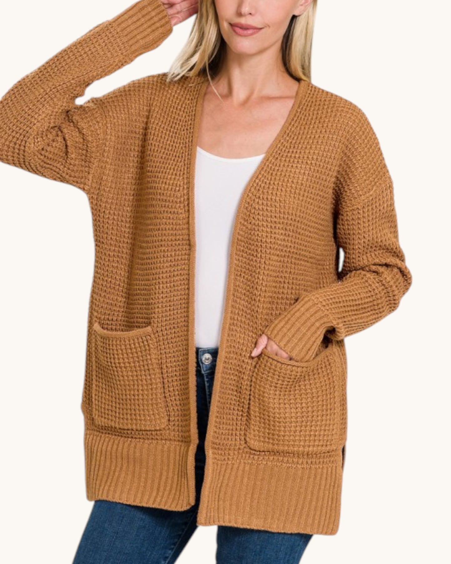 A woman wearing a chunky knit open-front cardigan in camel brown over a white tank top and blue jeans, shown against a plain light background.