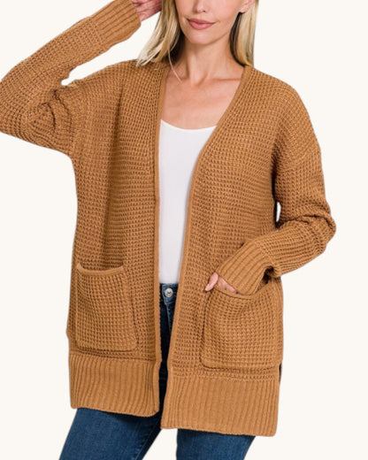 A woman wearing a chunky knit open-front cardigan in camel brown over a white tank top and blue jeans, shown against a plain light background.