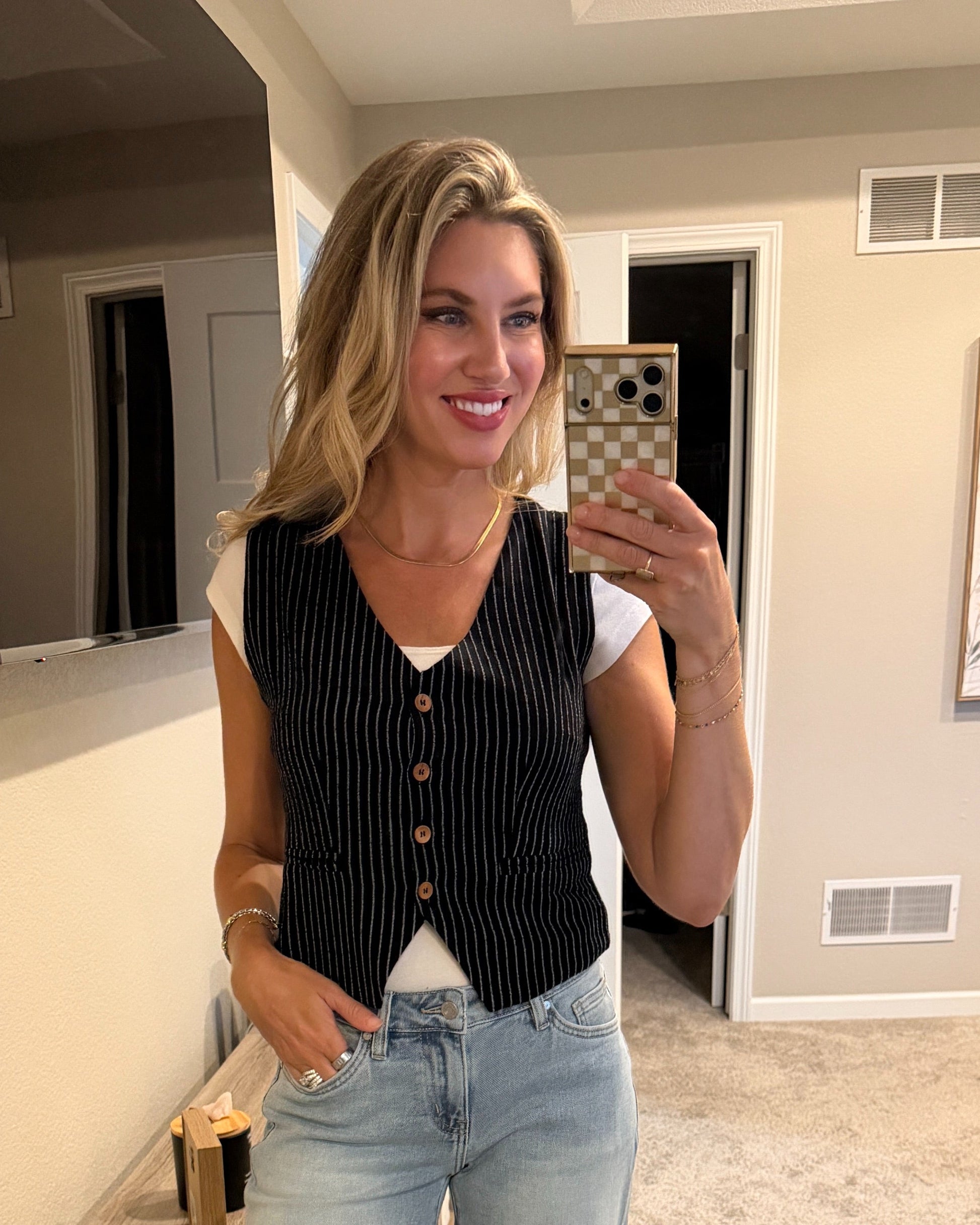 Woman wearing a black pinstripe vest with white tee shirt and light blue jeans taking a selfie.