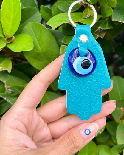 Blue keychain with an evil eye and hand design held by a hand against a green leafy background.