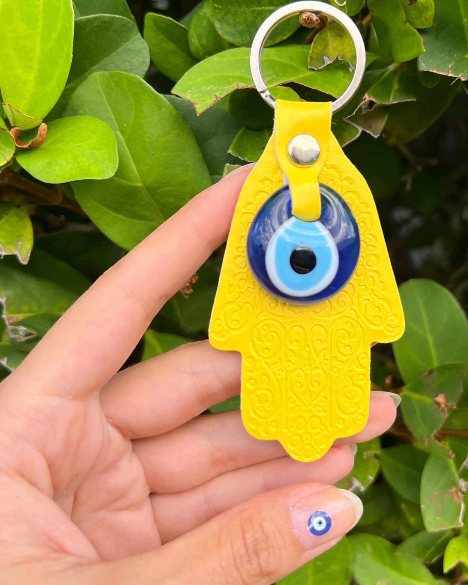 Yellow Hamsa keychain with an evil eye held by a hand against a green leafy background.