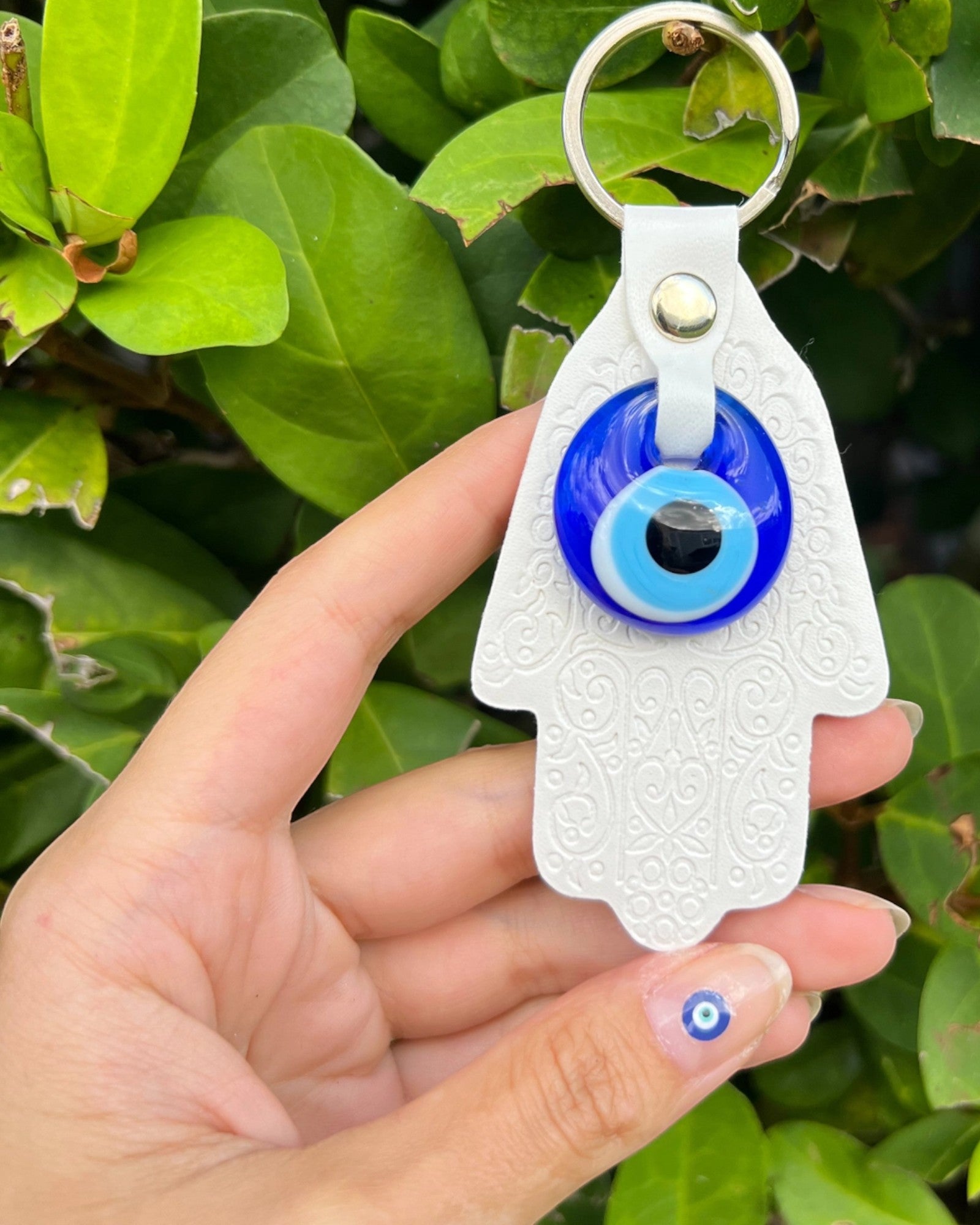 Hand holding a white Hamsa keychain with a blue evil eye against a green leafy background.