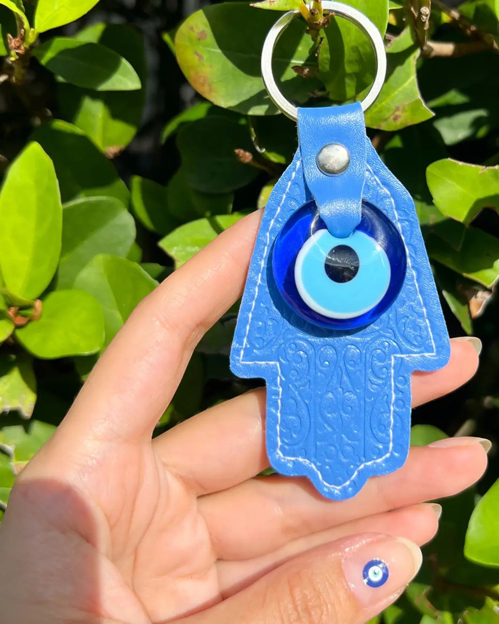 Blue Hamsa hand and evil eye keychain with a keyring held in front of green leaves.