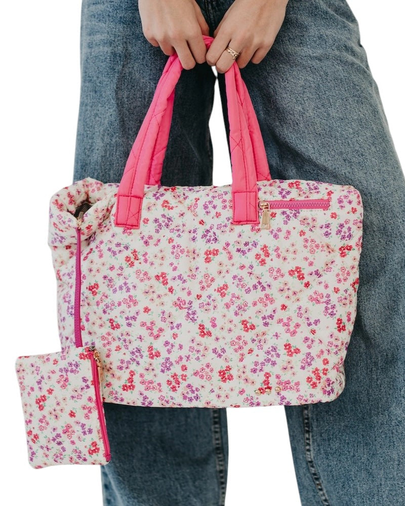 Floral-patterned quilted puffer tote bag with pink handles held by a person wearing jeans.