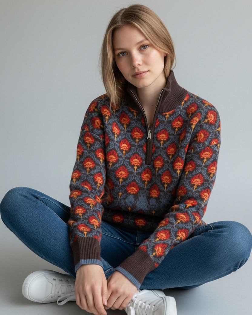 Woman wearing jeans and a patterned floral 1/2 zip brown and blue sweater with red and orange vintage floral accents on a grey background.