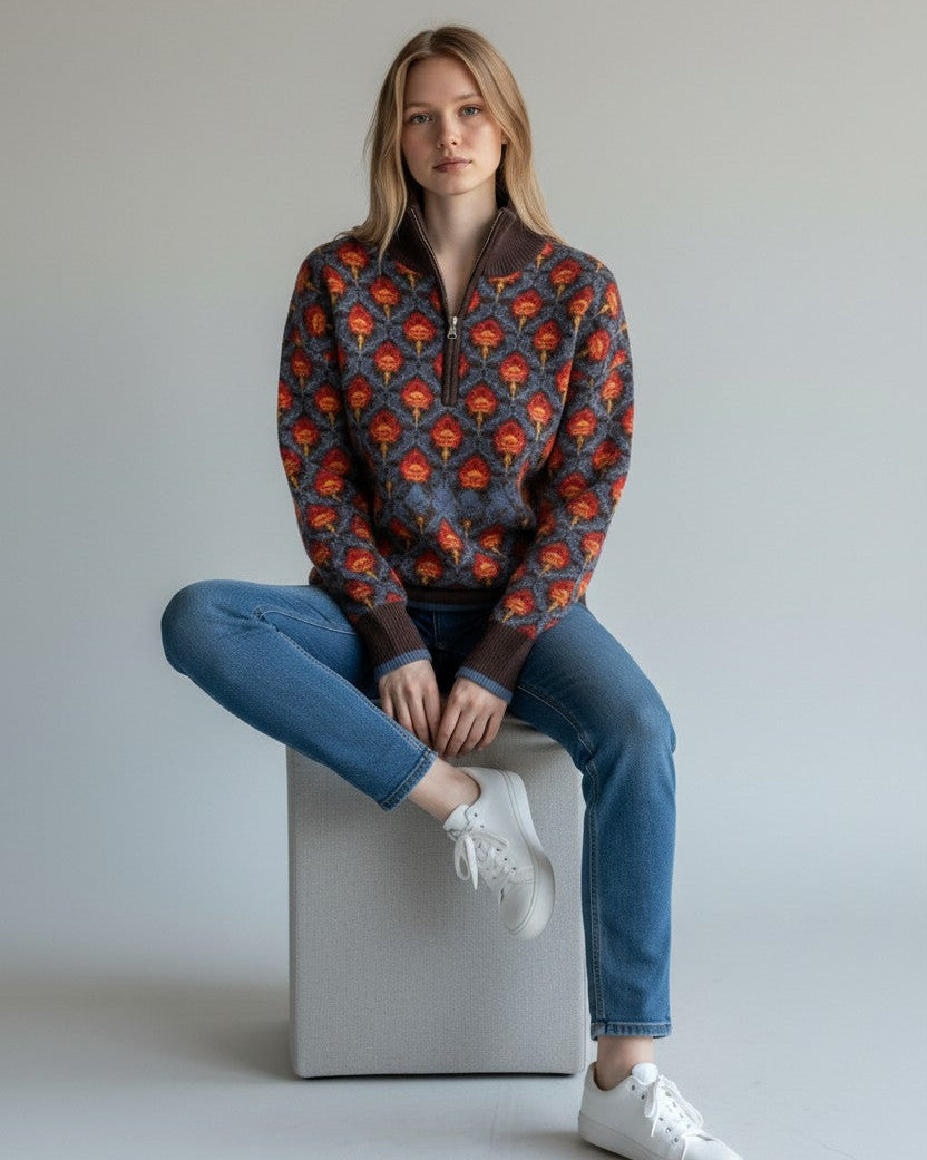 Woman sitting on a white cube wearing a patterned blue and brown floral 1/2 zip sweater and blue jeans.