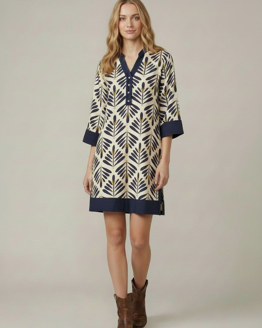 A woman modeling a short tunic dress with a navy blue and beige/gold geometric leaf pattern, featuring a V-neckline, buttoned placket, and three-quarter sleeves, paired with brown cowboy boots against a neutral studio background.