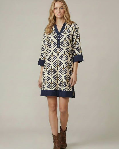 A woman modeling a short tunic dress with a navy blue and beige/gold geometric leaf pattern, featuring a V-neckline, buttoned placket, and three-quarter sleeves, paired with brown cowboy boots against a neutral studio background.