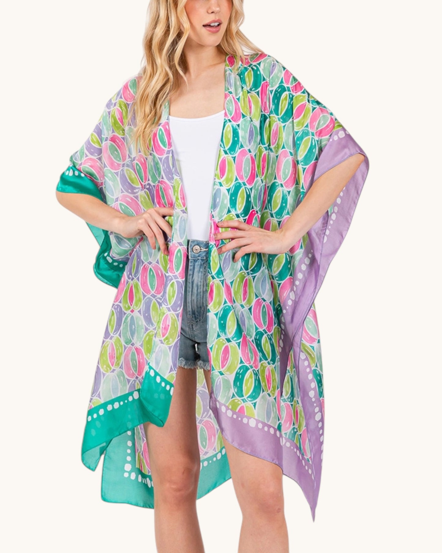Woman wearing a green and purple colorful patterned kimono over a white top and denim shorts on a white background.
