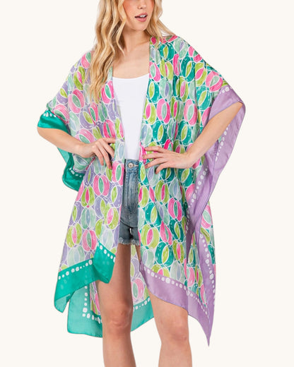 Woman wearing a green and purple colorful patterned kimono over a white top and denim shorts on a white background.
