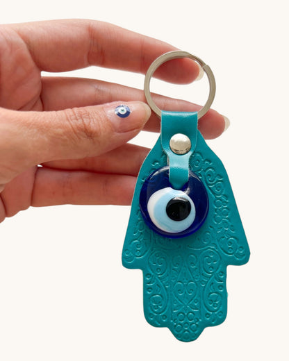 Turquoise Hamsa hand keychain with an evil eye charm held by a hand on a white background.
