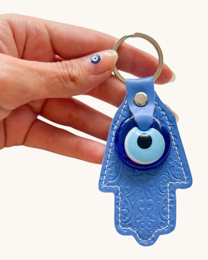 Blue Hamsa keychain with evil eye design held by a hand on a white background.