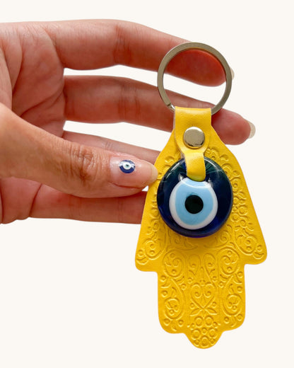 Yellow Hamsa keychain with an evil eye charm held by a hand on a white background.