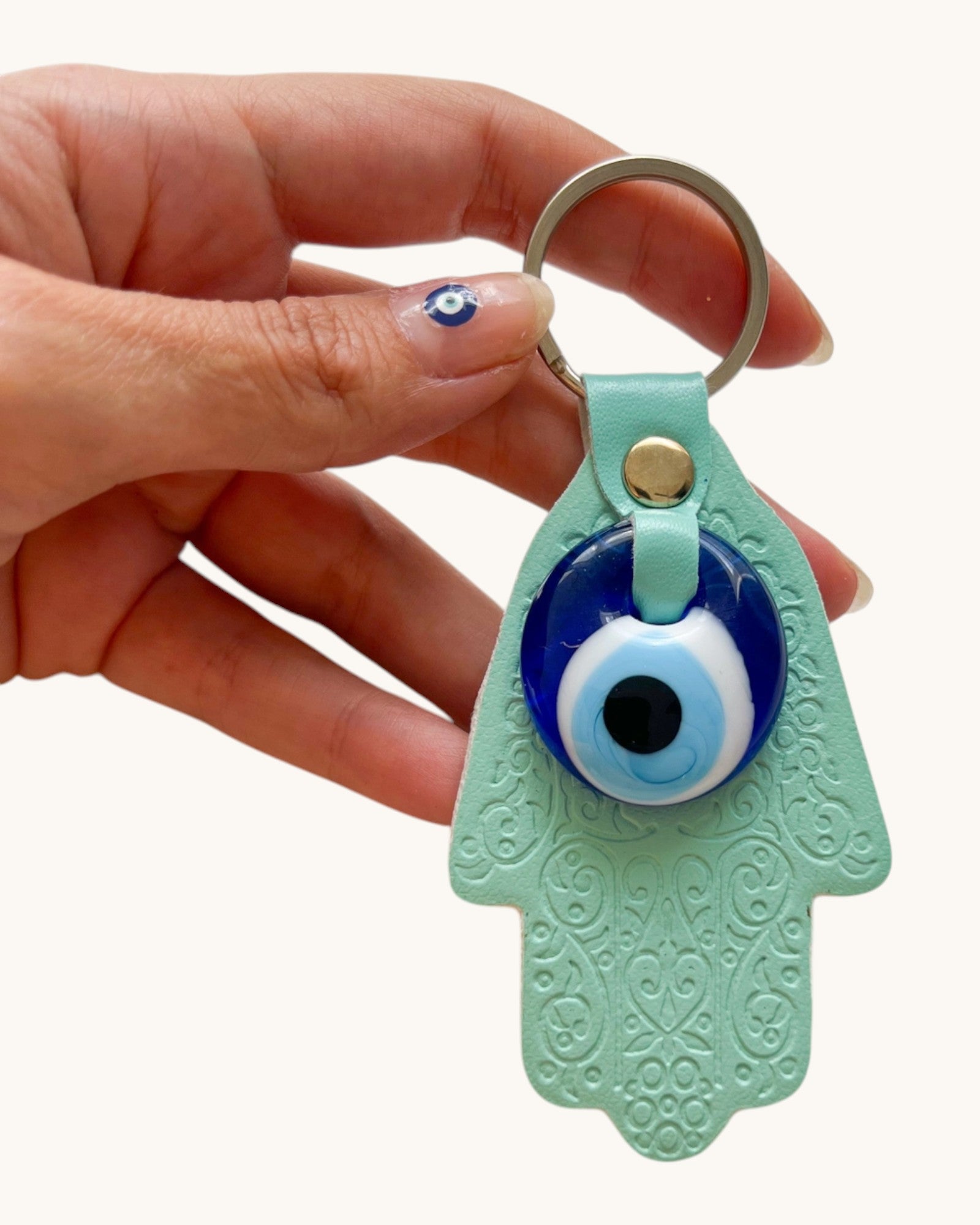 Hand holding a green Hamsa keychain with an evil eye design against a white background.