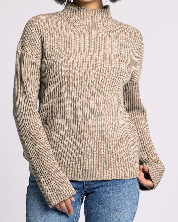 Woman wearing a beige ribbed sweater and blue jeans on a light gray background.