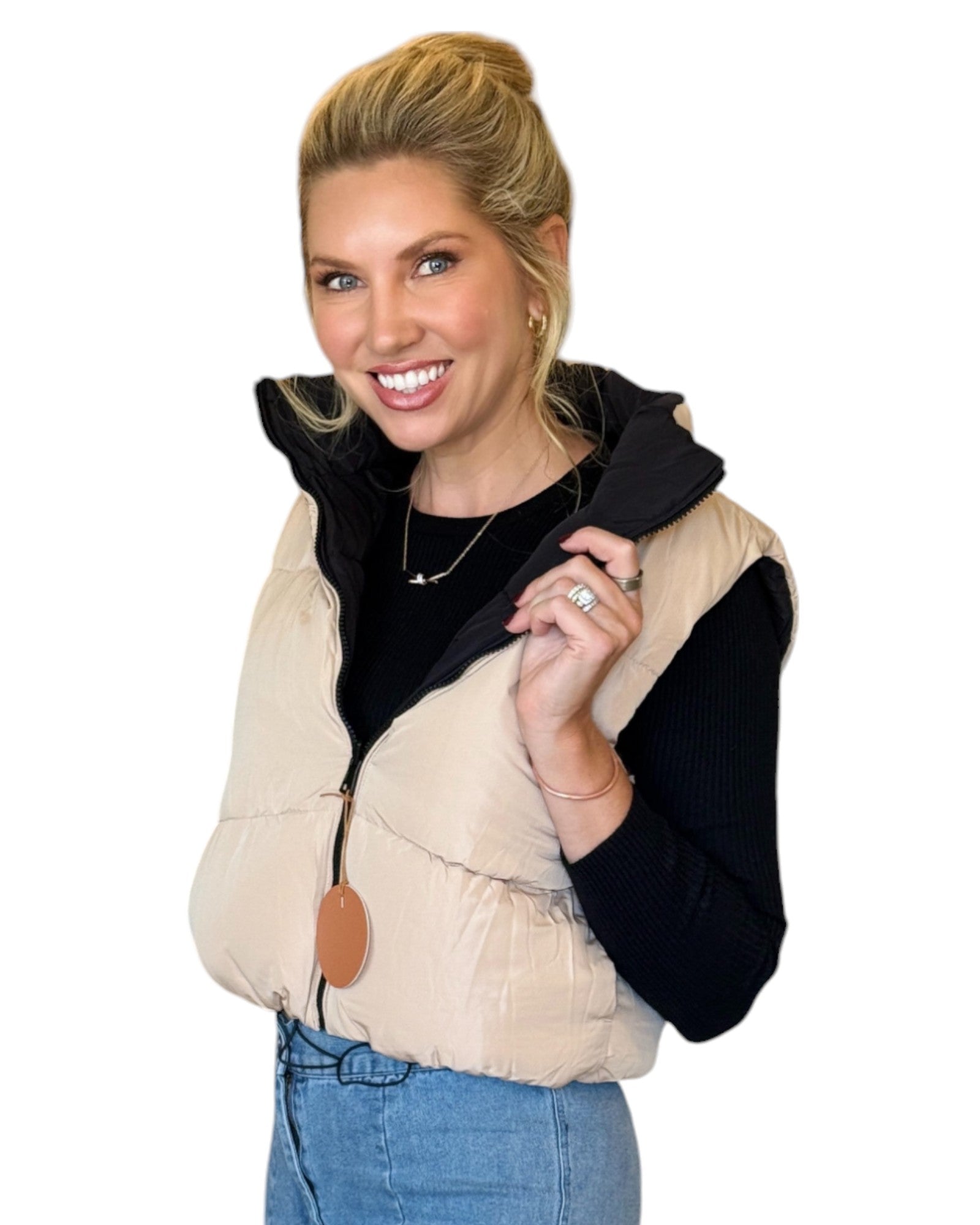 Woman wearing a beige and black reversible puffer vest with a zip-down front and funnel neck, paired with a black top and blue jeans.
