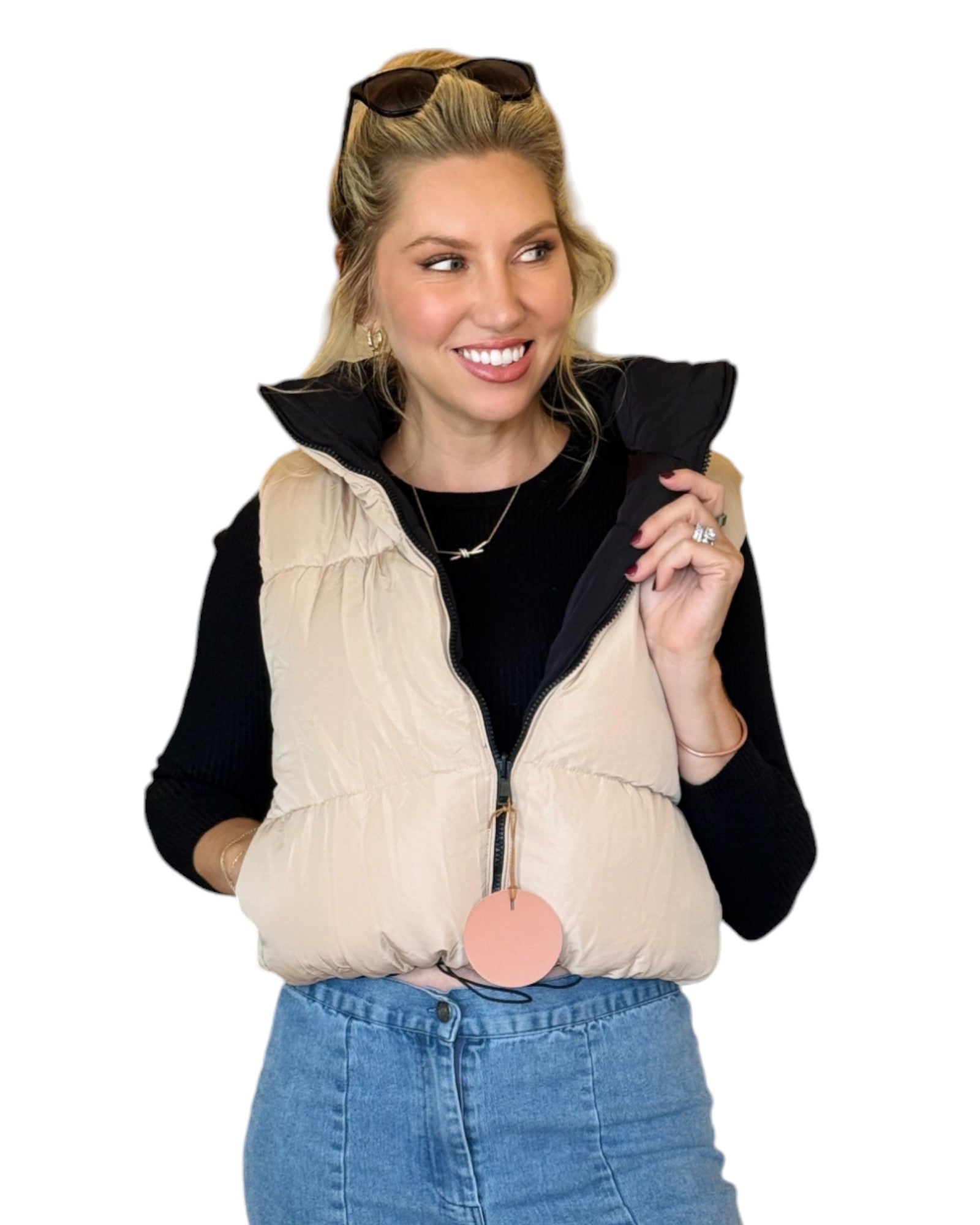 Woman wearing a beige and black reversible puffer vest with a zip-down front and funnel neck, paired with a black top and blue jeans.