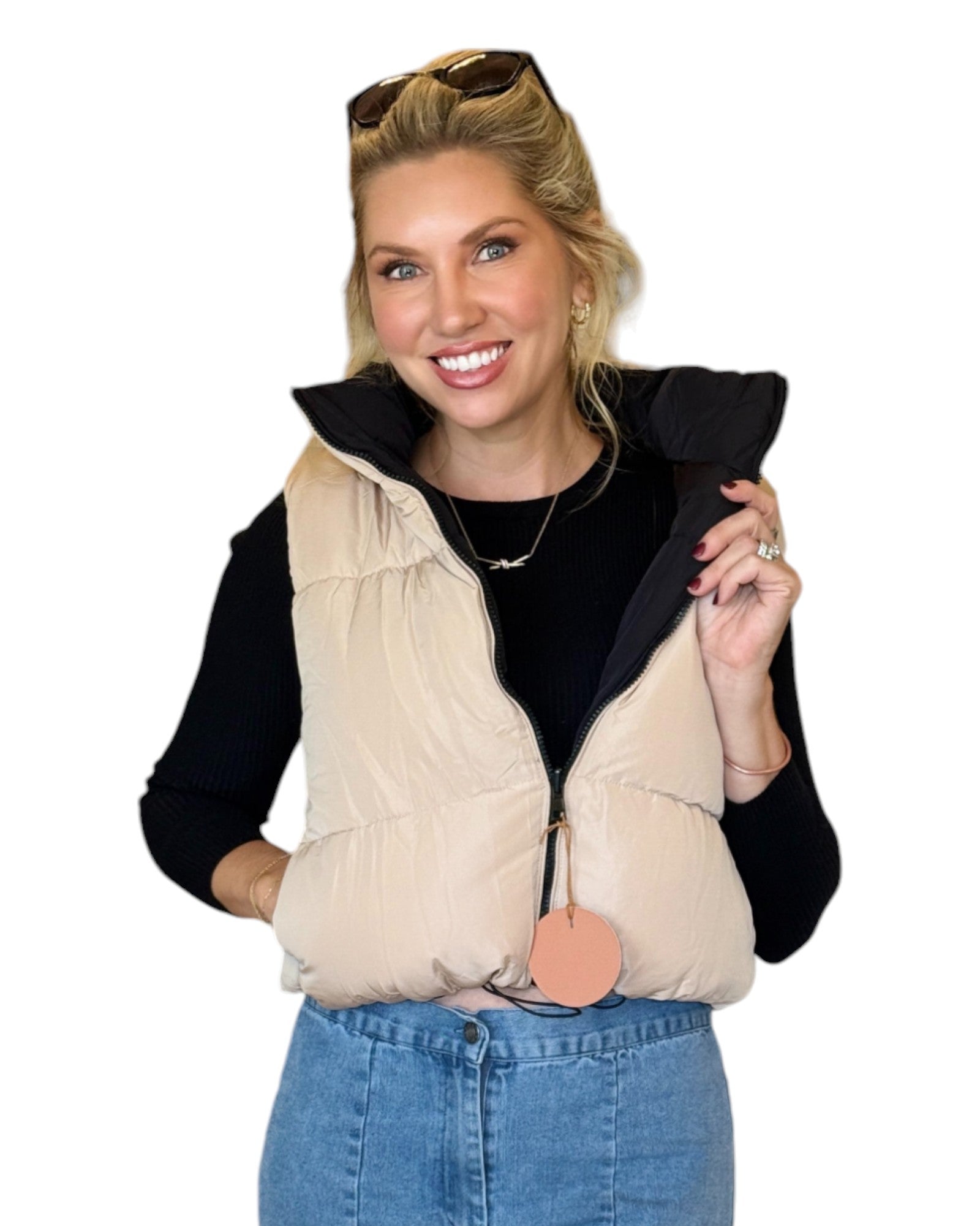 Woman wearing a beige and black reversible puffer vest with a zip-down front and funnel neck, paired with a black top and blue jeans.