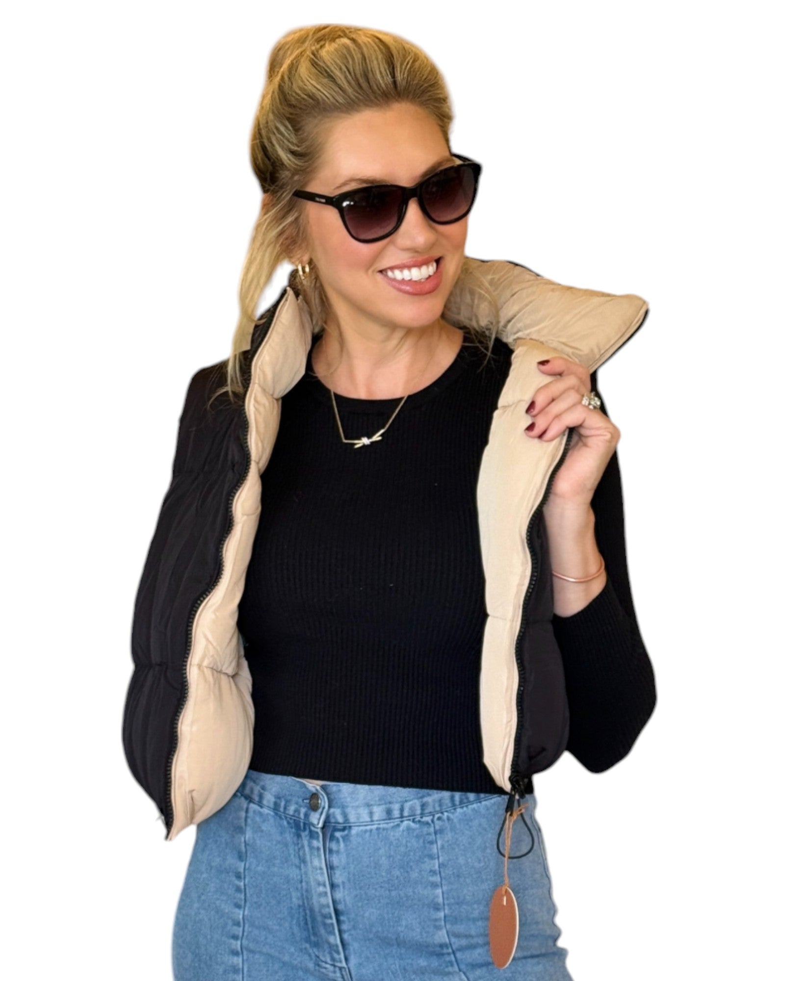 Woman wearing a beige and black reversible puffer vest with a zip-down front and funnel neck, paired with a black top and blue jeans.