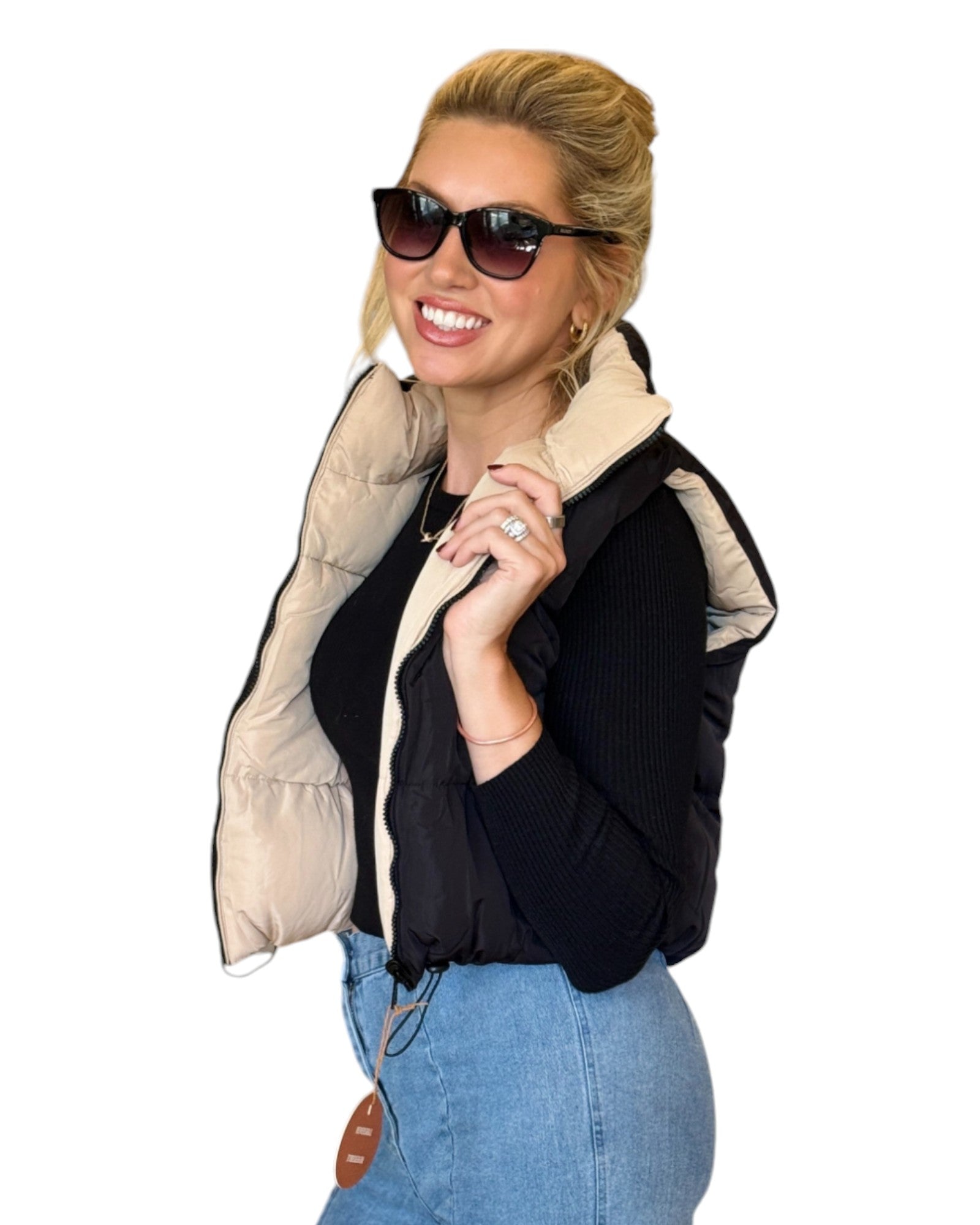 Woman wearing a beige and black reversible puffer vest with a zip-down front and funnel neck, paired with a black top and blue jeans.