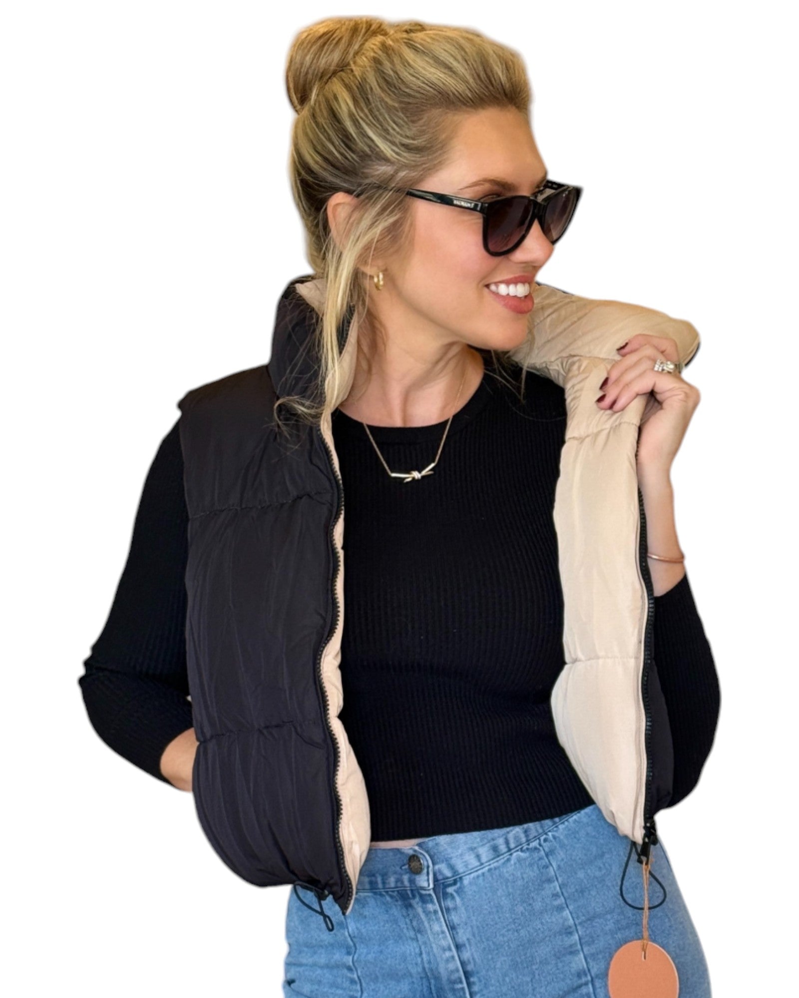 Woman wearing a beige and black reversible puffer vest with a zip-down front and funnel neck, paired with a black top and blue jeans.