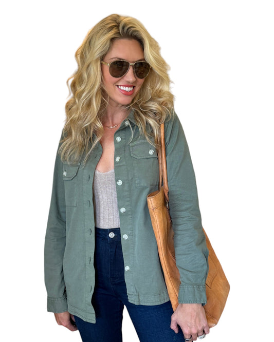 Woman wearing a sage green button-down shirt jacket layered over a ribbed tank top, paired with dark denim jeans. She accessorizes with aviator sunglasses and a tan leather tote bag, smiling with wavy blonde hair. The outfit combines casual, earthy tones for a chic and relaxed look.