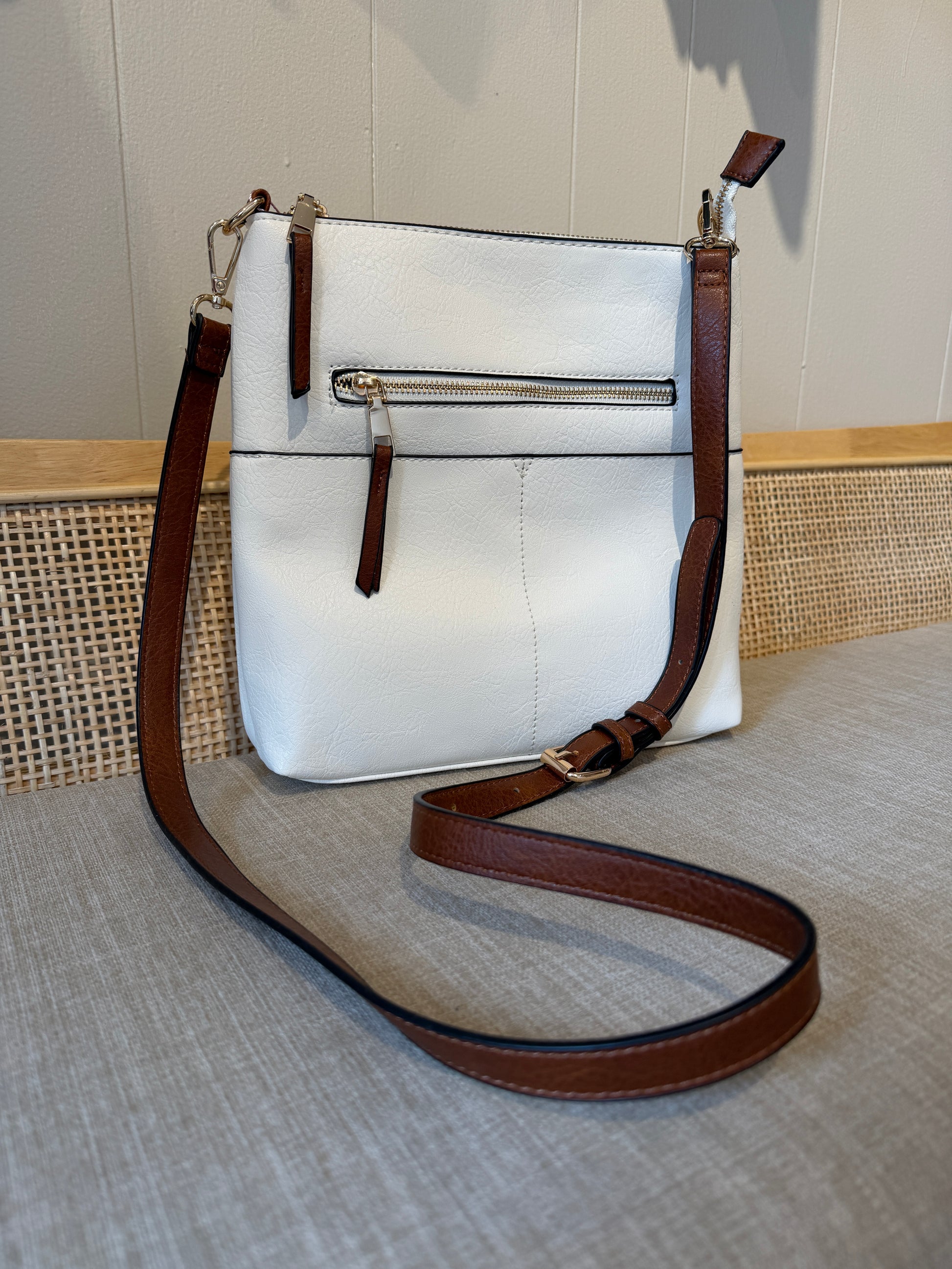 A white faux-leather crossbody bag with brown leather trim and strap, featuring a front zippered pocket, displayed on a textured beige surface.