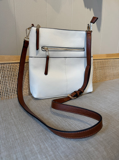 A white faux-leather crossbody bag with brown leather trim and strap, featuring a front zippered pocket, displayed on a textured beige surface.