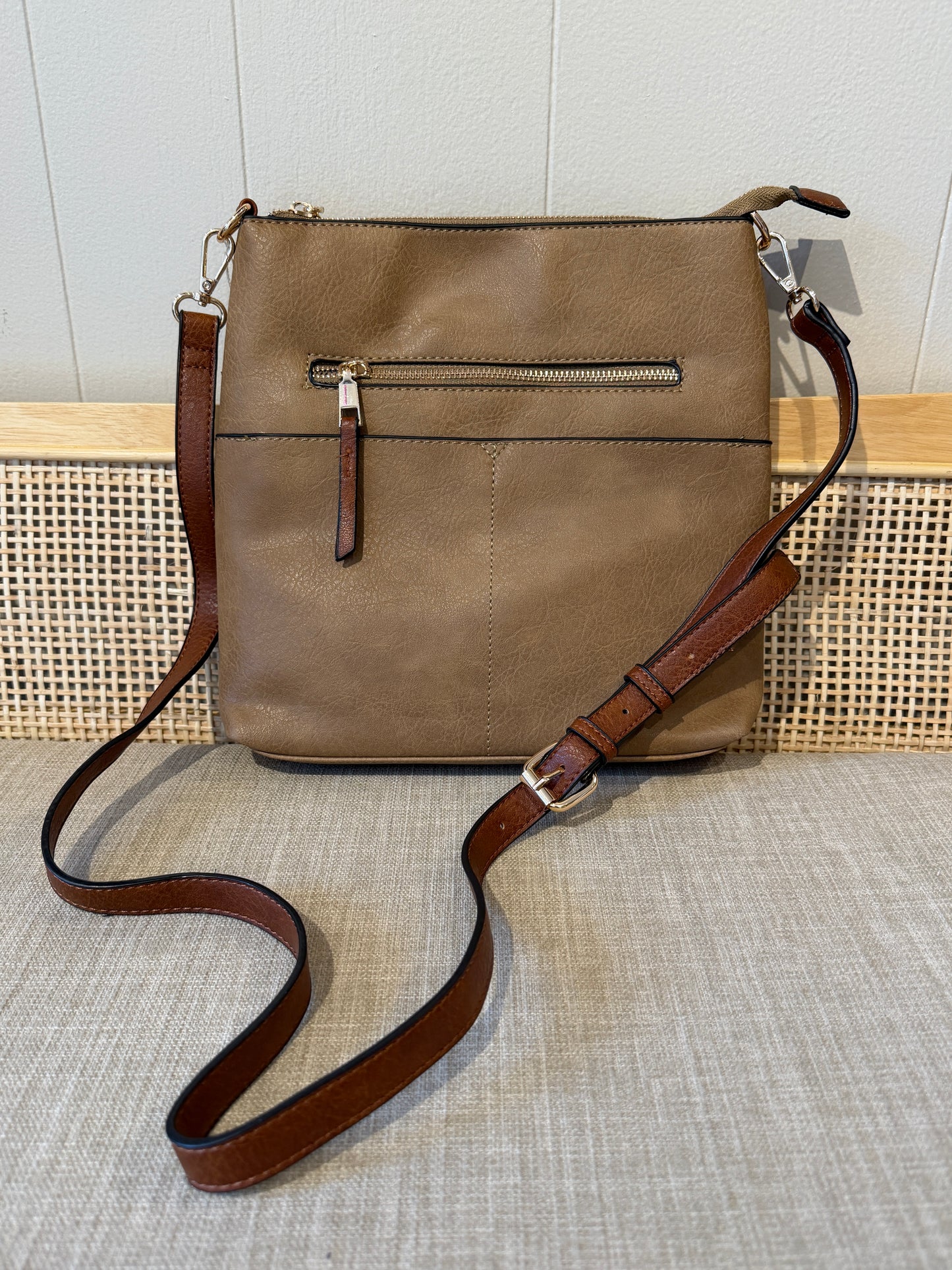 A taupe/mocha leather crossbody bag with a brown leather strap and gold-tone hardware, displayed on a textured beige fabric surface against a light wood and white paneled background.