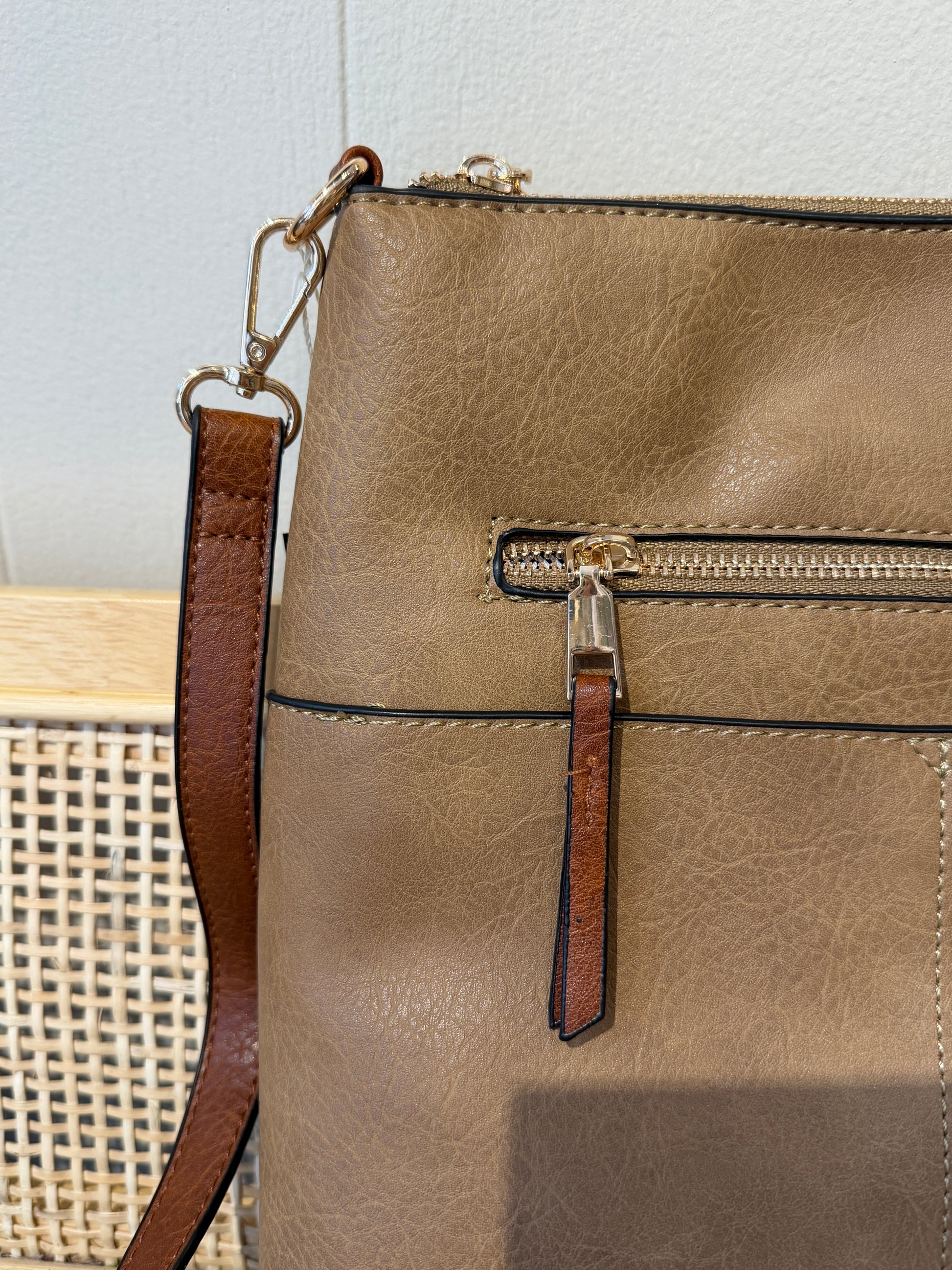 Close-up of a taupe faux-leather crossbody bag with a brown leather strap and gold-tone hardware, featuring a front zippered pocket with a leather pull tab.