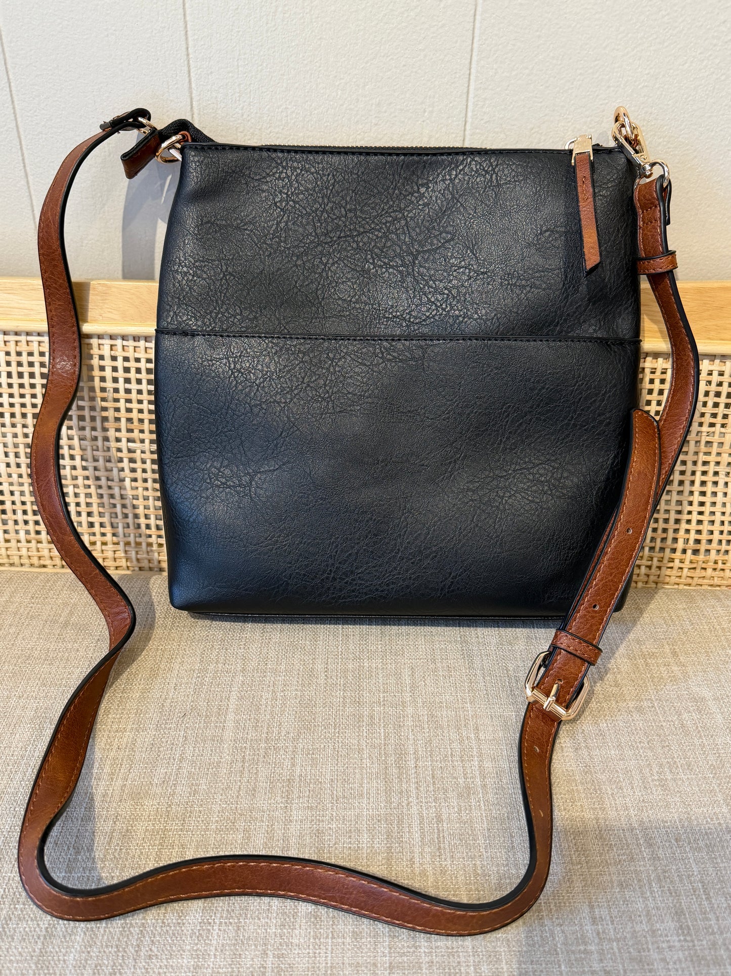 Black faux-leather crossbody bag with a brown leather strap and gold-tone hardware, displayed on a woven surface against a light wall.
