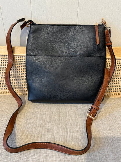 Black faux-leather crossbody bag with a brown leather strap and gold-tone hardware, displayed on a woven surface against a light wall.