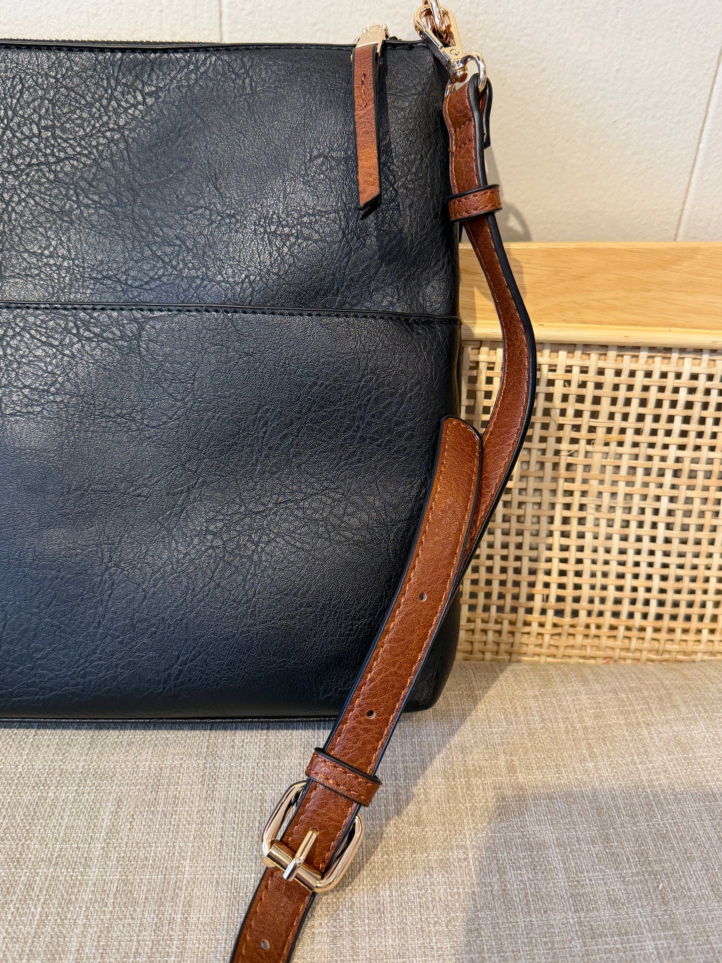 A close-up of a black leather shoulder bag with a brown leather strap and gold-tone buckle, resting on a woven beige surface.