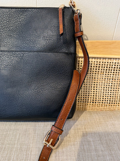 A close-up of a black leather shoulder bag with a brown leather strap and gold-tone buckle, resting on a woven beige surface.