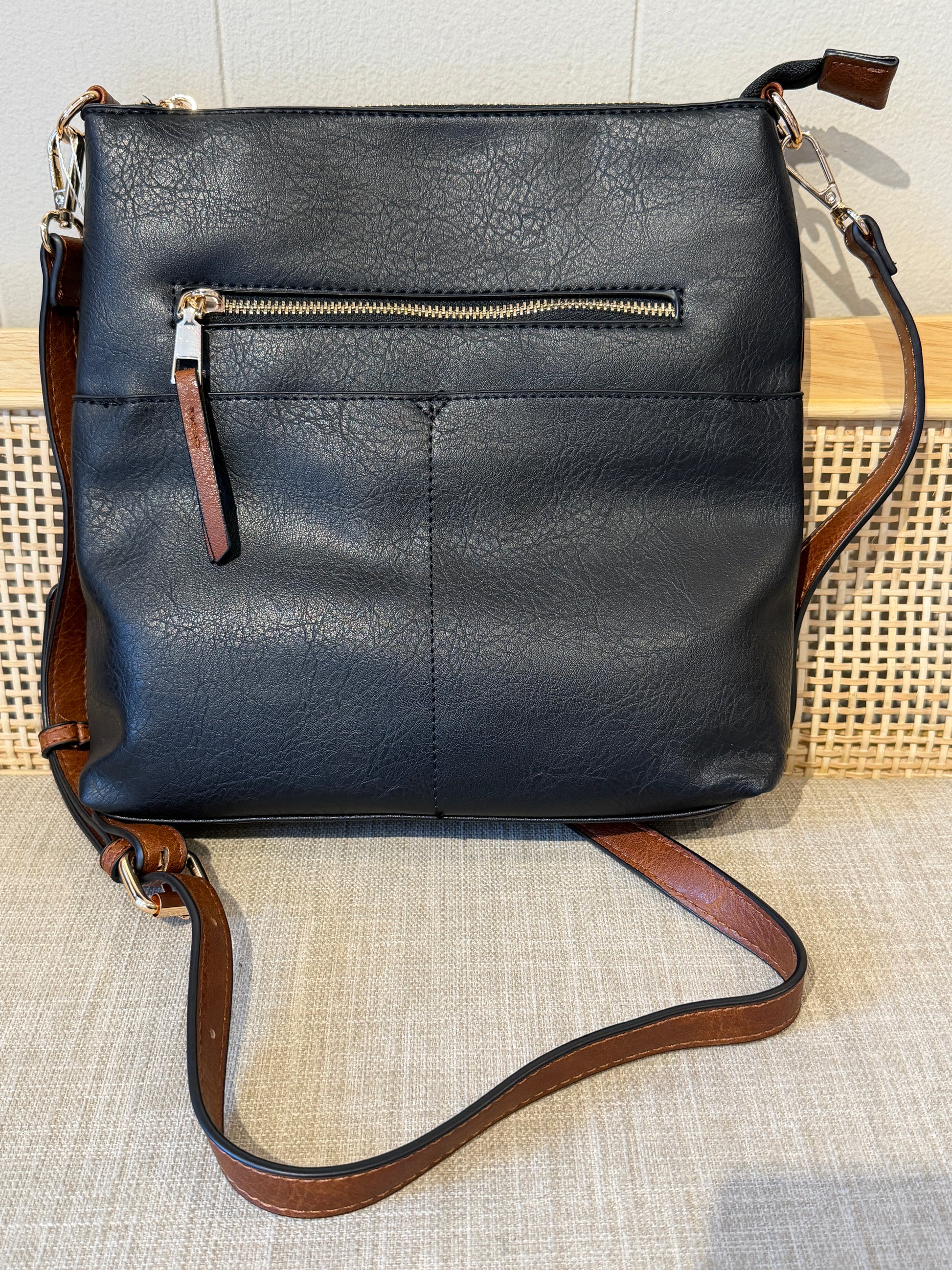 A black leather crossbody bag with a brown leather strap and trim, featuring a front zippered pocket, displayed on a textured beige surface.