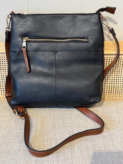 A black leather crossbody bag with a brown leather strap and trim, featuring a front zippered pocket, displayed on a textured beige surface.