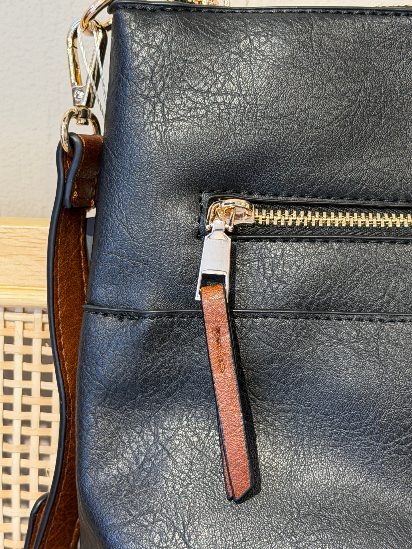 Close-up of a black pebbled leather handbag showing a gold-tone zipper with a brown leather pull tab and a matching brown shoulder strap.