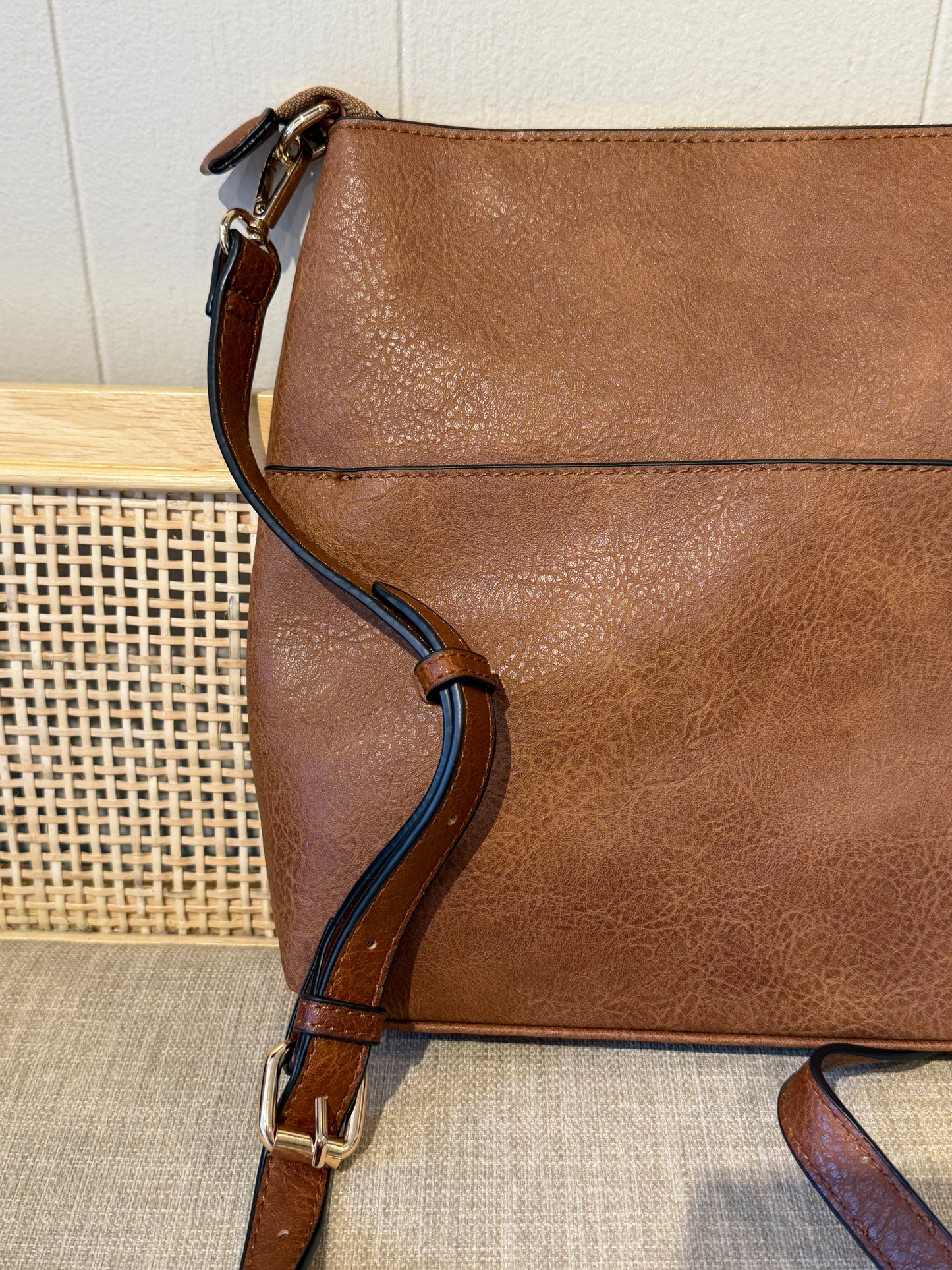 A close-up of a brown textured leather shoulder bag with a matching adjustable strap and gold-tone buckle, resting on a woven surface.