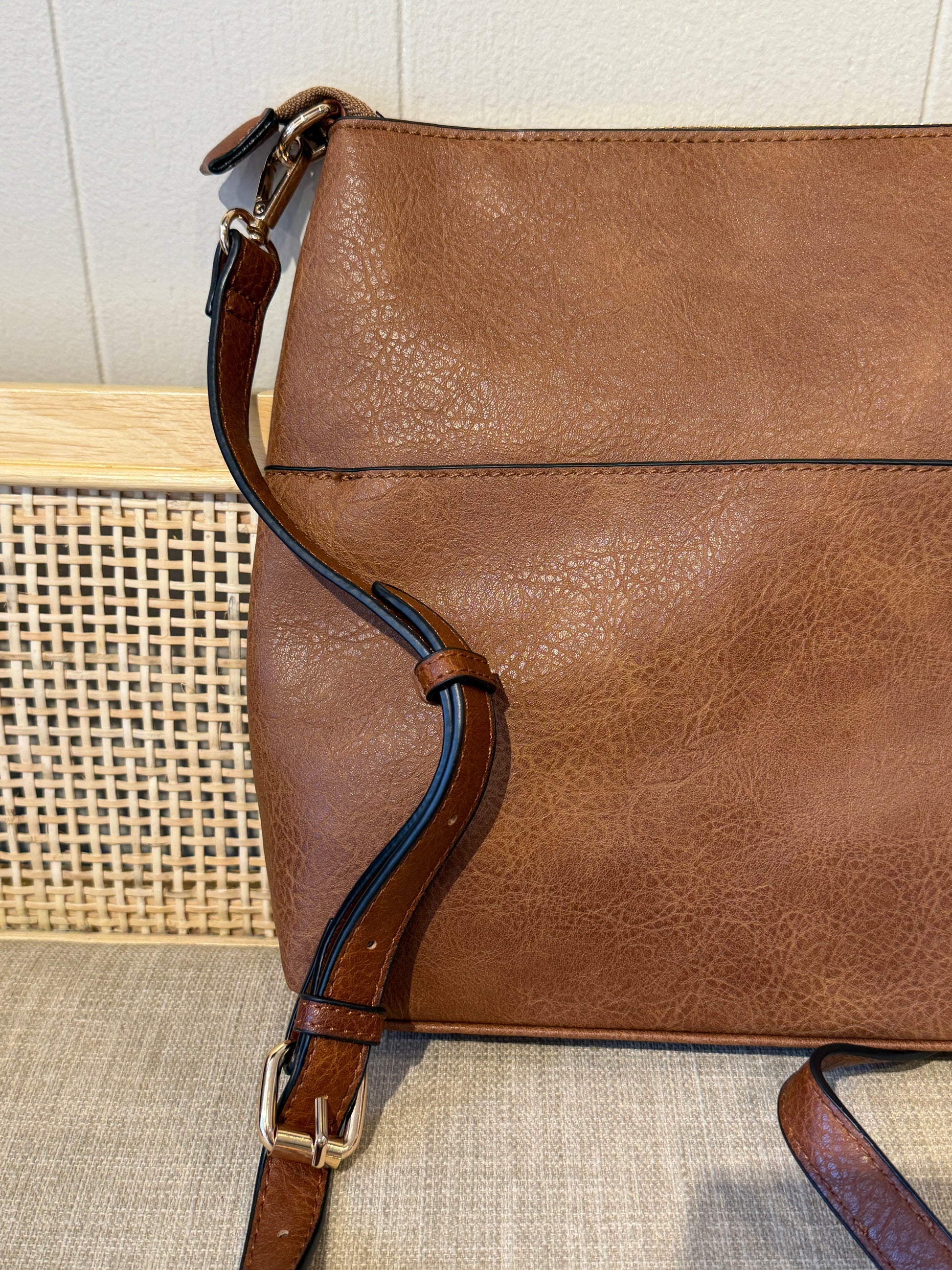 A close-up of a brown textured leather shoulder bag with a matching adjustable strap and gold-tone buckle, resting on a woven surface.