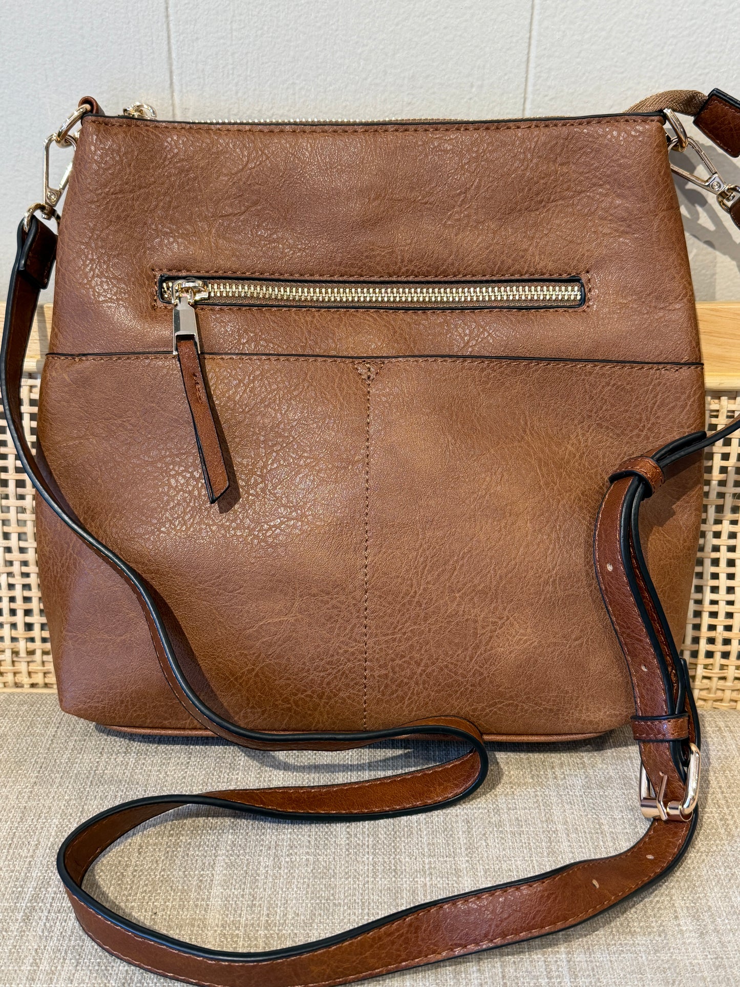 A brown faux-leather crossbody handbag with a back zippered pocket, shown on a textured beige surface against a light wall.