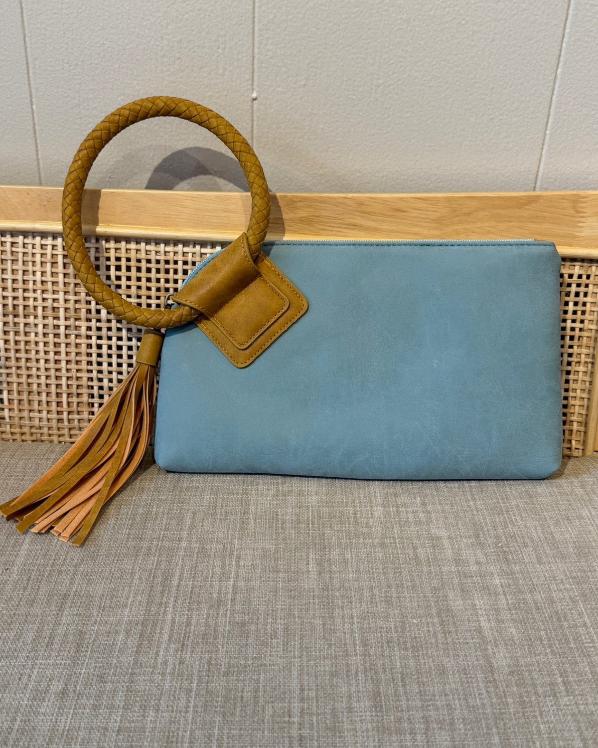 A light blue teal faux-leather clutch bag with a tan braided rope handle, leather tab, and tassel, displayed on a woven rattan bench against a light gray wall.