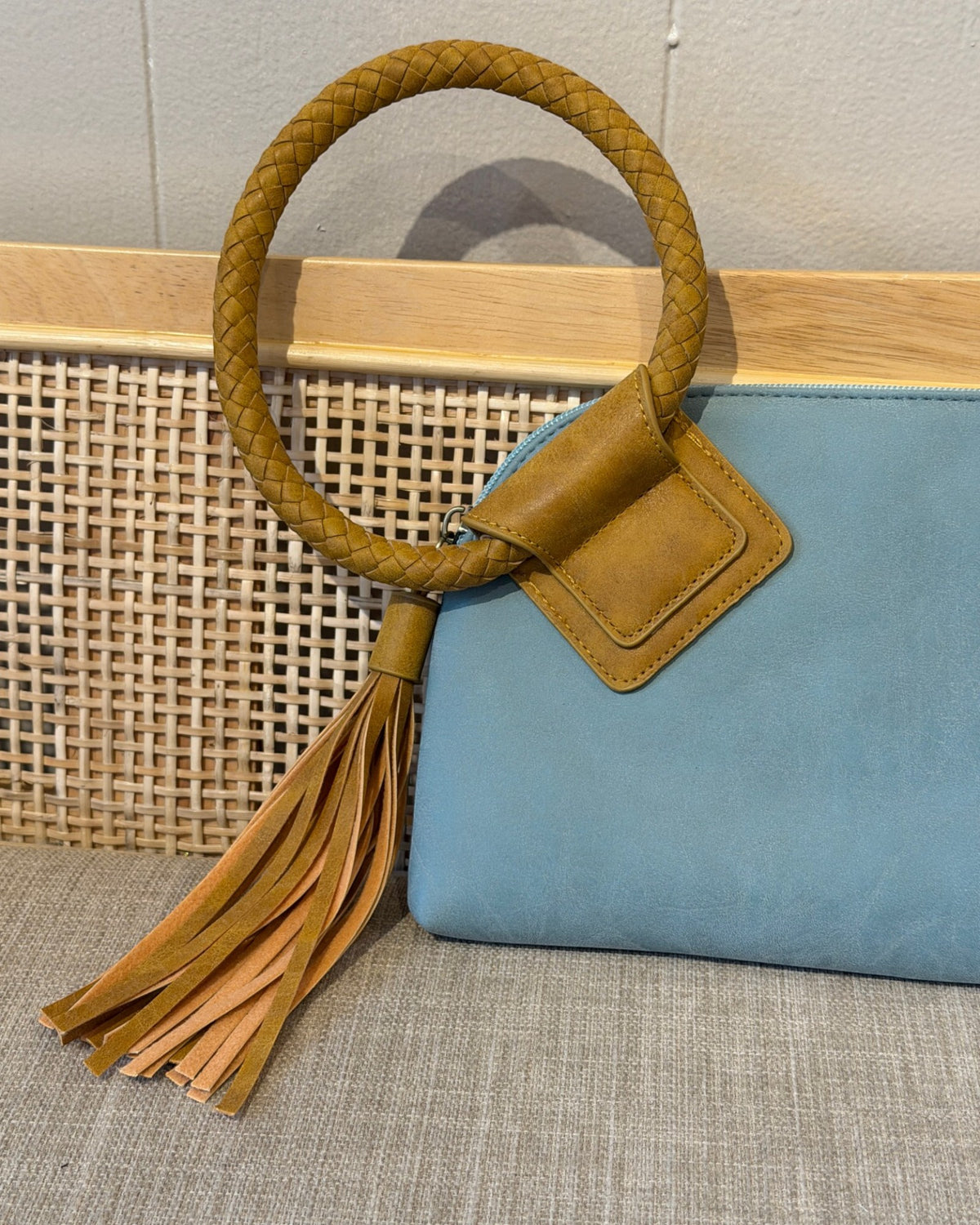 A light blue teal leather clutch bag with a thick braided tan handle and a matching tassel, displayed next to a woven cane panel.