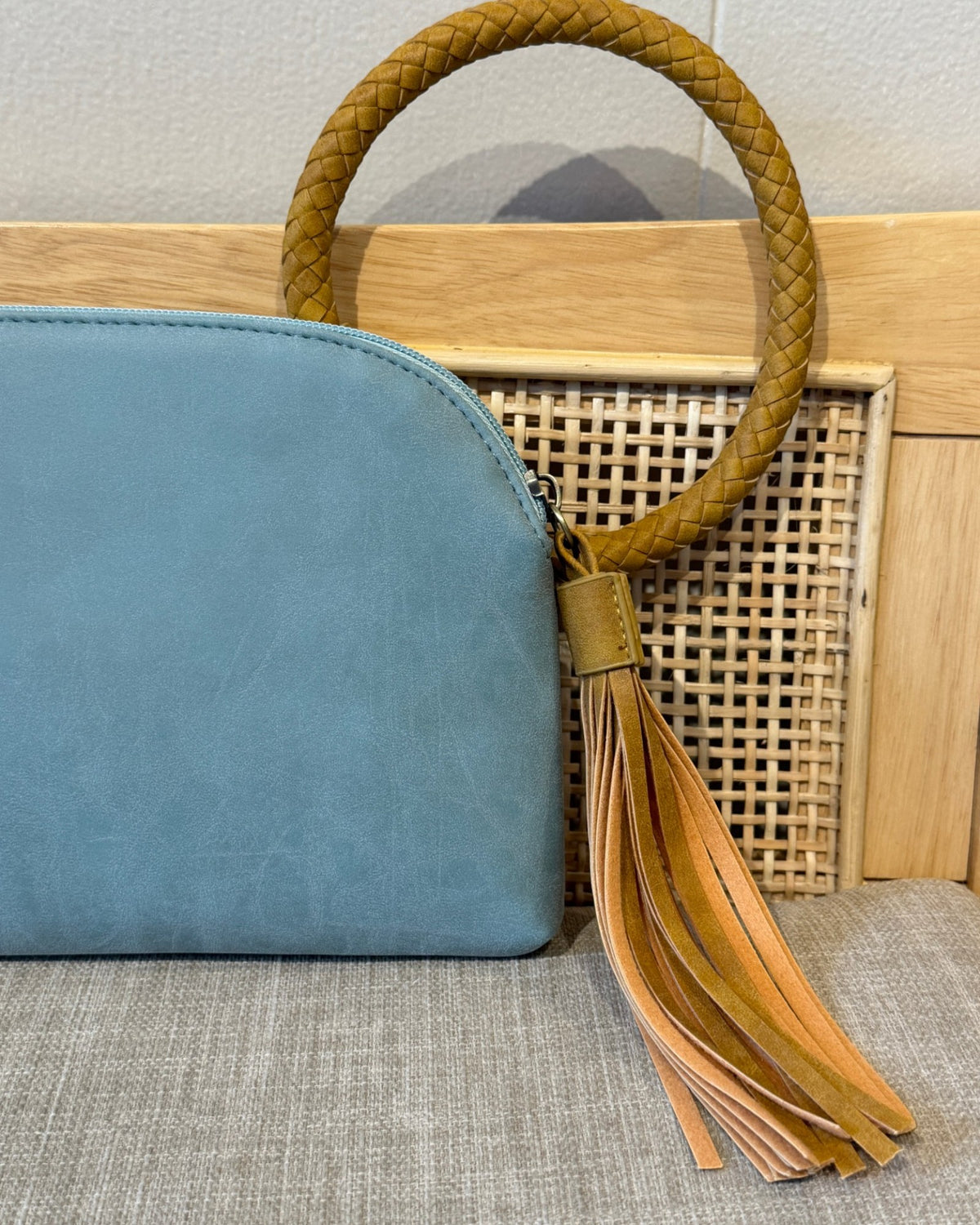 A light blue teal leather pouch bag with a curved top zipper, featuring a circular braided tan leather handle and a matching leather tassel charm, displayed next to a woven rattan box on a neutral textured surface.