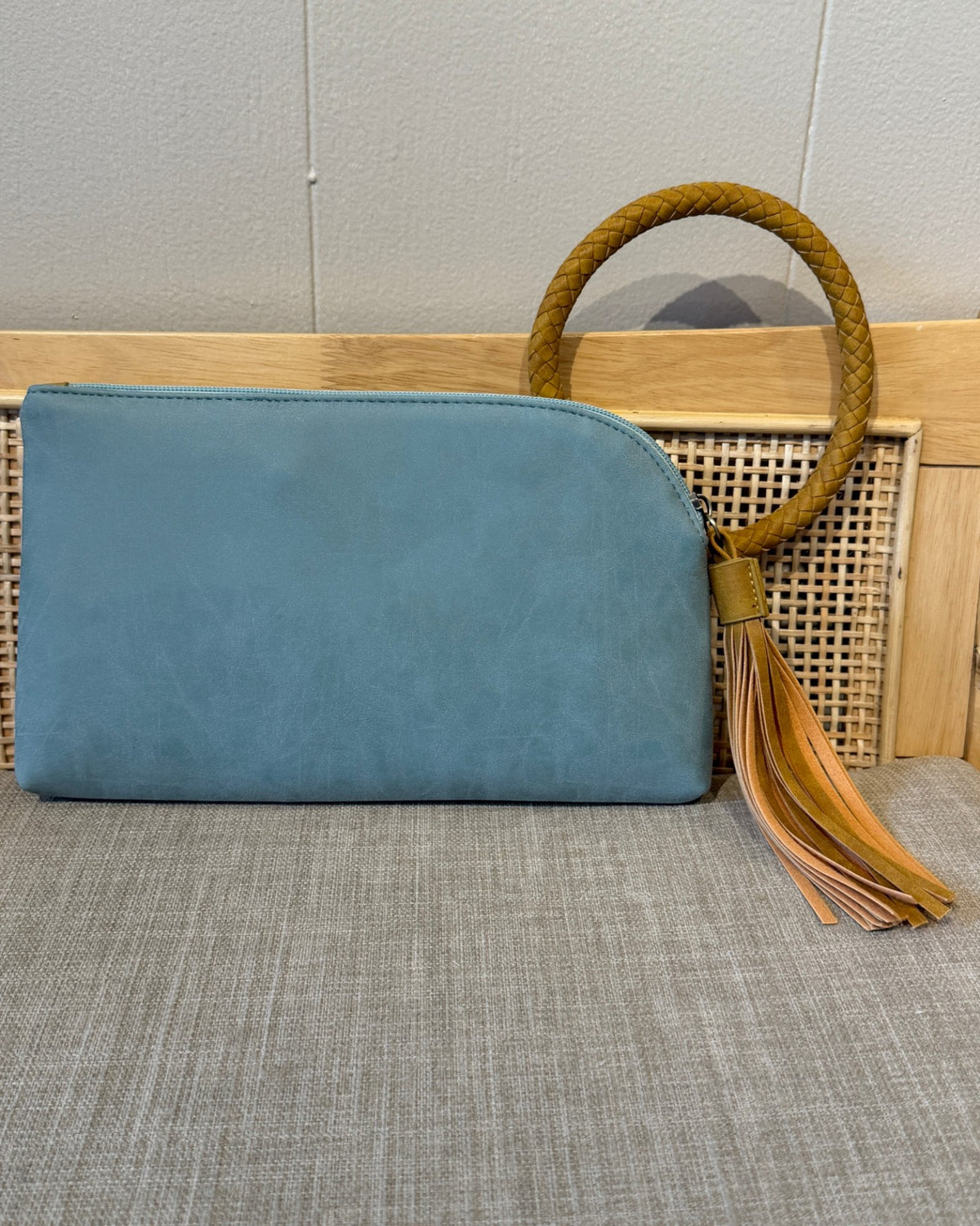 A light blue/teal faux-leather clutch bag with a curved top edge, a tan braided rope handle, and a matching leather tassel, displayed on a woven rattan bench against a light gray wall.