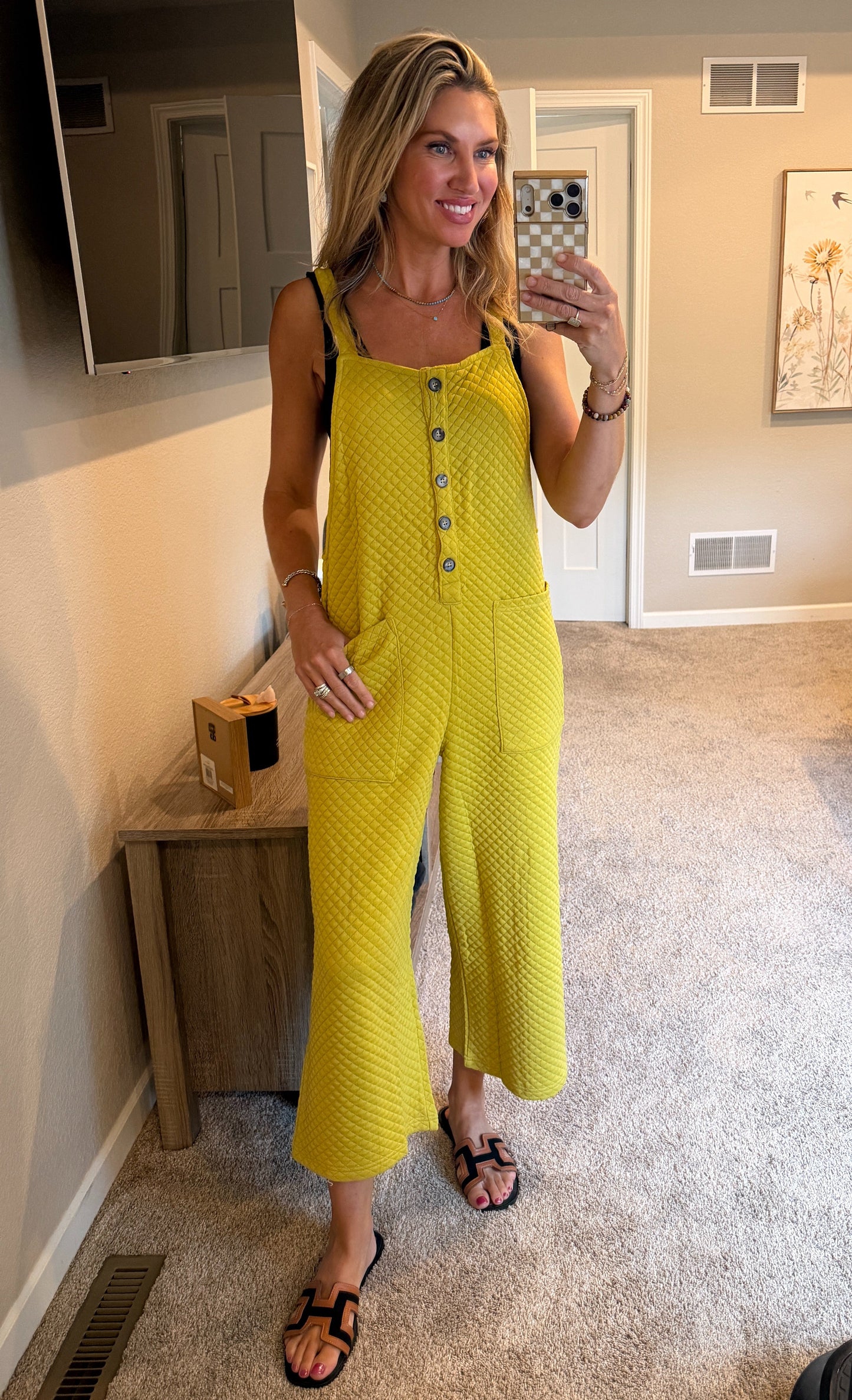 A woman takes a mirror selfie in a bedroom showing an avocado colored sleeveless textured jumpsuit with button details down the front and wide legs.