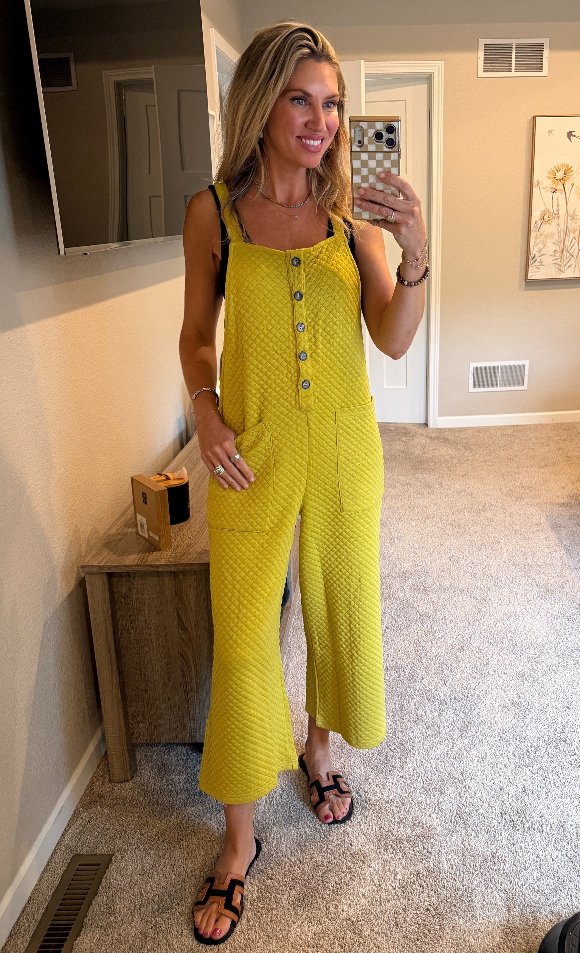 A woman takes a mirror selfie in a bedroom showing an avocado colored sleeveless textured jumpsuit with button details down the front and wide legs.