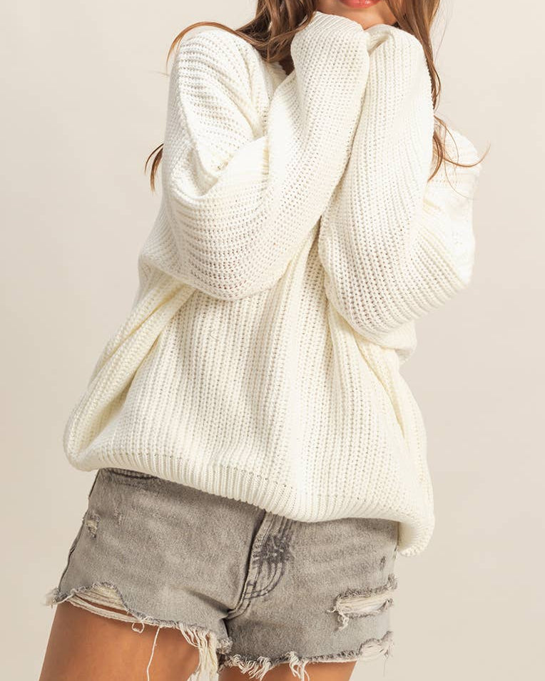 Woman wearing a white oversized sweater and denim shorts on a plain background.