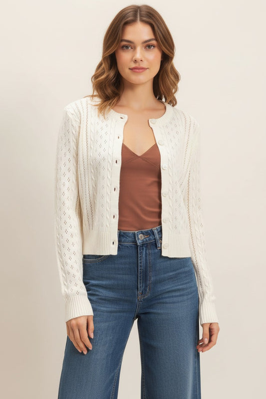 A woman modeling a cream cable-knit button-up cardigan over a brown camisole and blue jeans, photographed against a plain light beige studio background.
