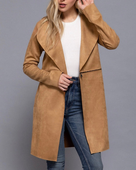Woman wearing a tan suede coat over a white shirt and blue jeans on a gray background.