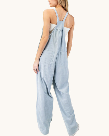 A woman shown from the back wearing a light blue corduroy jumpsuit with wide legs and shoulder straps, photographed against a plain off-white studio background.